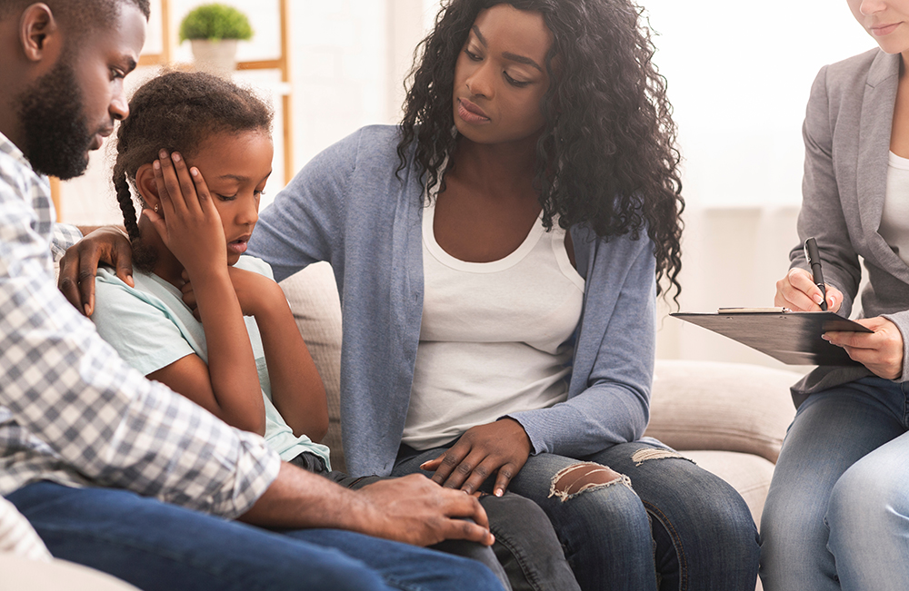 Childhood depression concept. Young black parents and their little daughter sitting at psychologist consultation, mother encouraging girl.